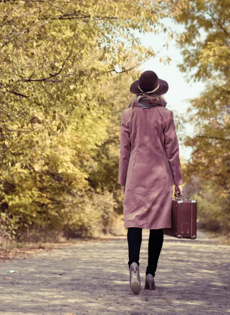 A traveller walking away down a tree-lined path in autumn, suitcase in hand