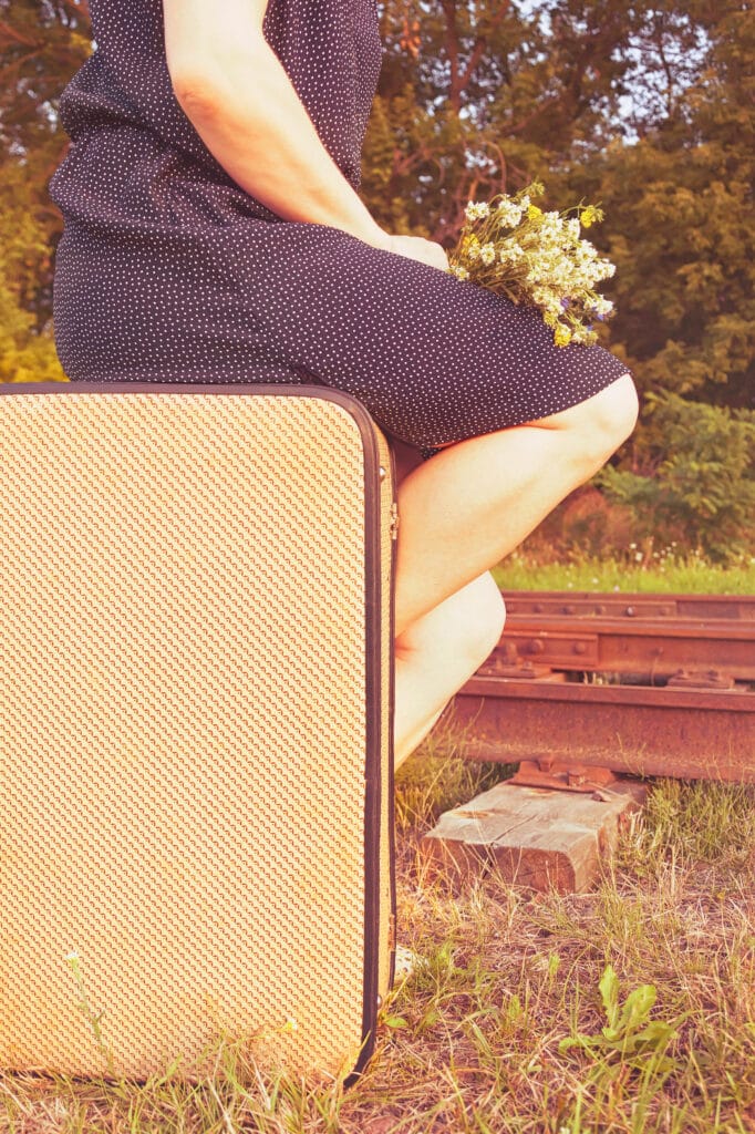 A traveller seated on a vintage suitcase beside railway tracks, holding wildflowers